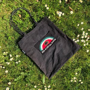 Puede incluir: Un bolso de tela negro con un parche rectangular bordado. El parche muestra una rodaja de sandía estilizada en rojo, blanco y verde con una semilla negra central y el texto blanco ALL EYES ON GAZA cosido debajo de la fruta.