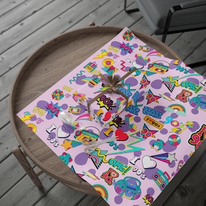 May include: A rectangular gift box wrapped in light pink paper featuring a repeating pattern of 1990s-themed icons including boomboxes, Rubik's cubes, rainbows, lightning bolts, and the text 90's, OMG, and STAY COOL, tied with a brown ribbon.