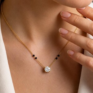 May include: A gold chain necklace featuring a single round brilliant-cut clear gemstone pendant at the center, flanked by two small black beads on each side of the chain, creating a delicate and refined piece of jewelry for the neckline.