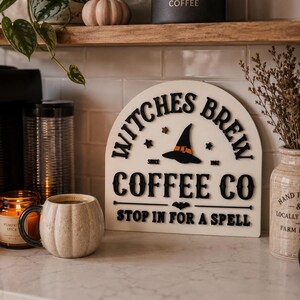 May include: An arched off-white wooden sign with black 3D lettering that reads Witches Brew Coffee Co, featuring a black witch hat with an orange band, small stars, a bat icon, and the text Stop In For A Spell, with the date Since 1982.