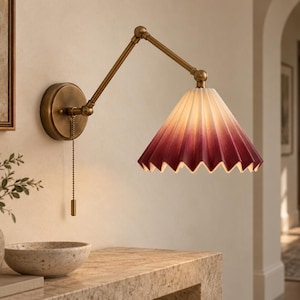 May include: A wall-mounted swing arm lamp with an antique brass finish and a pull chain switch. The lamp features a conical, pleated fabric shade that transitions from a light cream colour at the top to a deep, saturated burgundy at the bottom edge.
