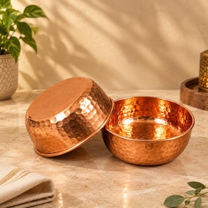 May include: Two round bowls crafted from polished copper, each featuring a distinct hammered texture across the exterior surface. One bowl is positioned upright, while the other rests tilted against it, showcasing the reflective metallic finish.