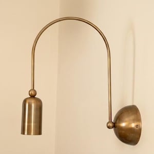 May include: A wall-mounted lamp with a brushed brass finish. The fixture features a semi-circular arched arm connecting a dome-shaped wall plate to a cylindrical downward-facing light socket, accented by a small spherical connector piece.