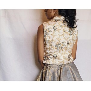 Puede incluir: Un chaleco sin mangas de tela color crema con un intrincado patrón de bordado floral en dorado y azul oscuro, combinado con una falda gris con un sutil diseño geométrico de jacquard. El chaleco tiene cuello alto y una silueta estructurada.
