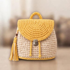 May include: A crochet backpack featuring a cream-colored body and a bright yellow flap, handle, and base trim. The bag includes a silver-toned metal push-lock closure, two yellow drawstring cords, and a decorative yellow tassel with wooden beads.