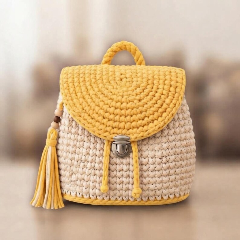 May include: A crochet backpack featuring a cream-colored body and a bright yellow flap, handle, and base trim. The bag includes a silver-toned metal push-lock closure, two yellow drawstring cords, and a decorative yellow tassel with wooden beads.