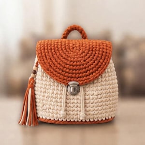 May include: A crochet backpack featuring a cream-colored body and a burnt orange flap. The bag includes a top handle, a silver-toned metal push-lock closure, and a decorative tassel made of cream and orange cord hanging from the side.