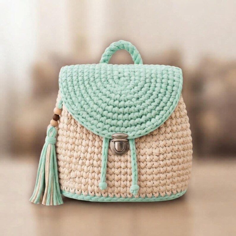 May include: A crochet backpack featuring a beige main body and a mint green flap. The bag includes a top handle, a silver-toned metal push-lock closure, and a decorative side tassel made of mint green and beige yarn with small wooden beads.