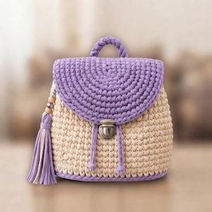 May include: A crochet backpack featuring a beige main body and a lavender flap with a top handle. The bag includes a silver-toned metal push-lock closure, two lavender drawstring cords, and a decorative lavender tassel with wooden beads on the side.