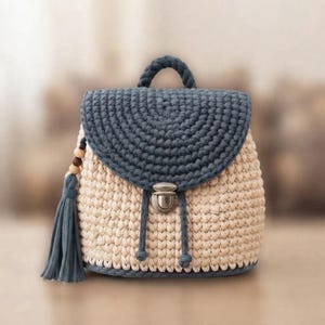 May include: A crochet backpack featuring a cream-colored body and a dark blue flap. The bag includes a top handle, a metal push-lock closure, and a dark blue tassel accent decorated with wooden beads hanging from the side of the main compartment.