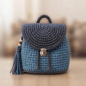 May include: A crochet backpack featuring a light blue body and a dark gray flap. The bag includes a top handle, a silver-toned metal push-lock closure, and a decorative side tassel with wooden beads attached to a dark gray cord.