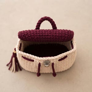 May include: A small crochet backpack featuring a cream-colored body and a dark burgundy flap, handle, and drawstring cords. The bag includes a silver-toned metal clasp closure on the front and a decorative tassel with cream and burgundy strands.