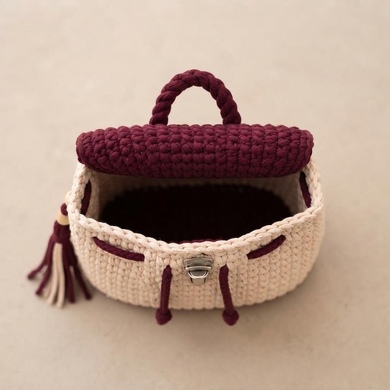 May include: A small crochet backpack featuring a cream-colored body and a dark burgundy flap, handle, and drawstring cords. The bag includes a silver-toned metal clasp closure on the front and a decorative tassel with cream and burgundy strands.