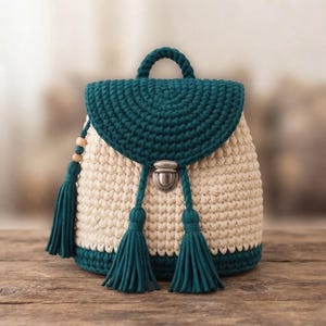 May include: A handcrafted crochet backpack featuring a cream-colored body with a dark teal flap, base, and handle. The bag includes a silver-toned metal push-lock closure and three dark teal tassels, with one tassel accented by small wooden beads.