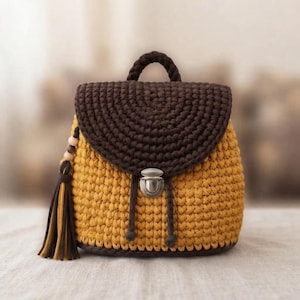 May include: A crochet backpack featuring a mustard yellow body and a dark brown flap. The bag includes a top handle, a silver-toned metal push-lock closure, and a decorative tassel on the side with wooden beads and matching yellow and brown strands.