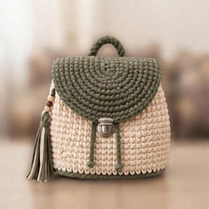 May include: A crochet backpack featuring a cream-colored body and a dark olive green flap. The bag includes a top handle, a metallic silver push-lock closure, and a decorative tassel on the side with wooden beads and matching green and cream yarn.