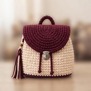 May include: A crochet backpack featuring a cream-colored body and a dark burgundy flap. The bag includes a top handle, a silver-toned metal push-lock closure, and a decorative side tassel made of burgundy and cream yarn with wooden beads.