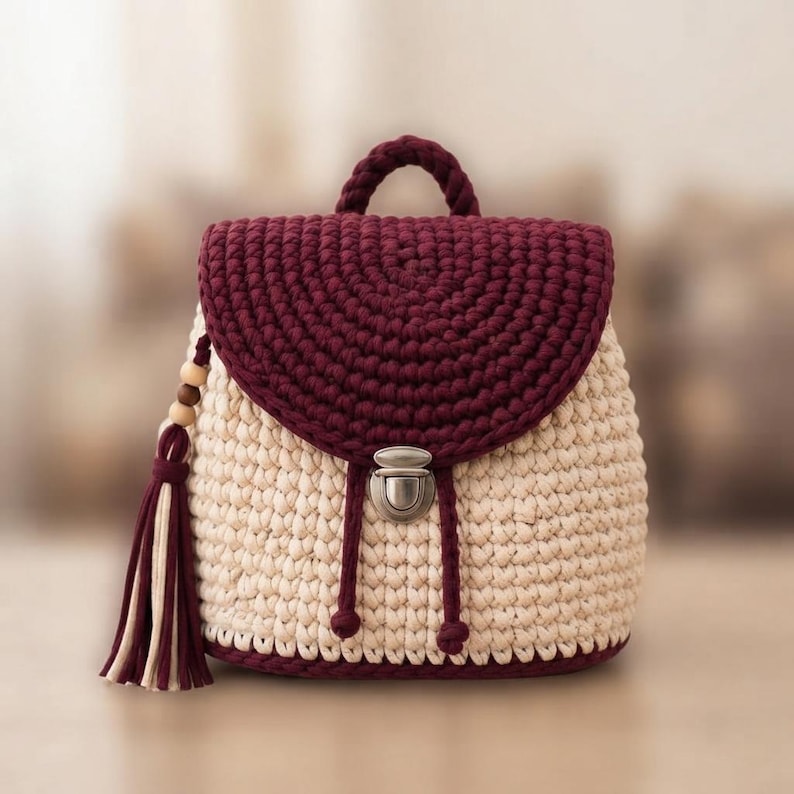 May include: A crochet backpack featuring a cream-colored body and a dark burgundy flap. The bag includes a top handle, a silver-toned metal push-lock closure, and a decorative side tassel made of burgundy and cream yarn with wooden beads.