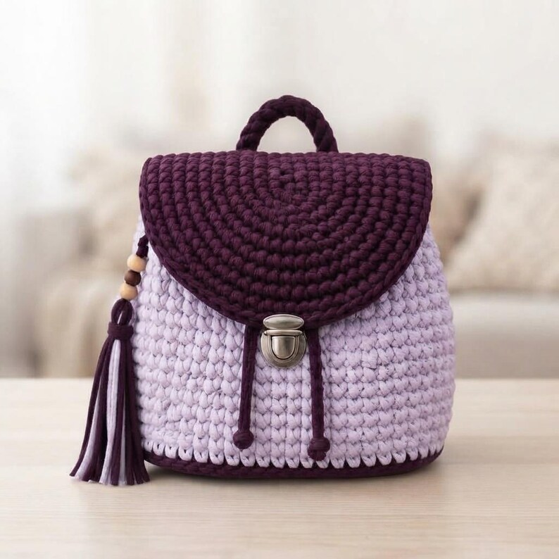 May include: A crochet backpack featuring a light lavender body and a dark purple flap. The bag includes a top handle, a metallic silver push-lock closure, and a decorative side tassel adorned with three wooden beads in varying shades of brown.