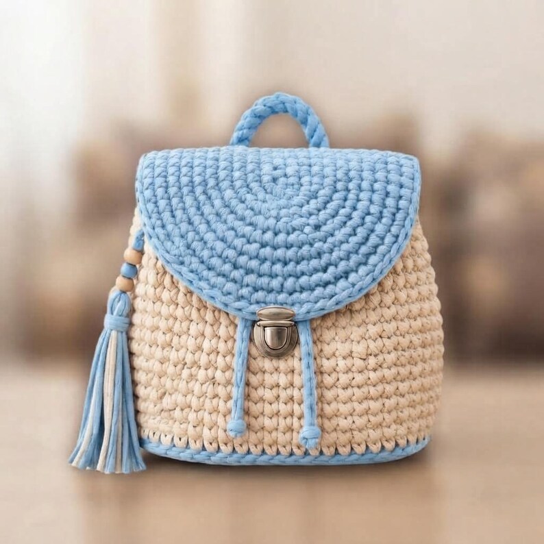 May include: A crochet backpack featuring a cream-colored main body and a light blue flap, handle, and base trim. The bag includes a silver-toned metal push-lock closure and a decorative tassel on the side with light blue and cream yarn strands.