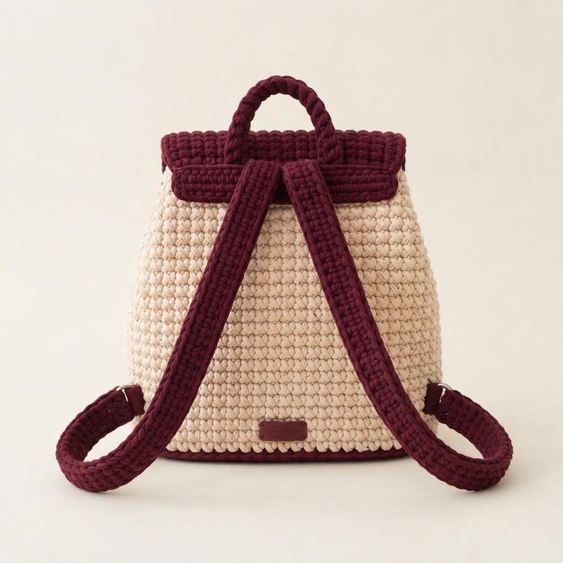 May include: A crochet backpack featuring a cream-colored main body and a dark burgundy flap, top handle, and adjustable shoulder straps. The bag is constructed with a dense, textured stitch pattern and includes a small rectangular patch at the base.