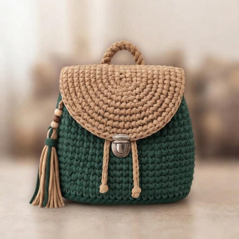 May include: A crochet backpack featuring a dark green body and a tan flap. The bag includes a top handle, a silver-toned metal push-lock closure, and a decorative tassel on the side with wooden beads. The construction consists of thick yarn.
