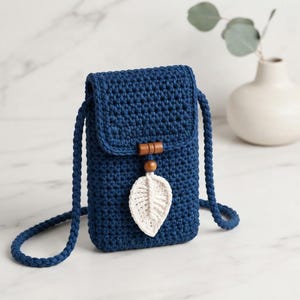 May include: A dark blue crochet crossbody bag featuring a rectangular flap with a magnetic closure. The bag is adorned with a decorative wooden bead and a white macrame leaf charm hanging from the center. It includes a matching braided strap.