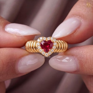 Può includere: Un anello in oro con una pietra preziosa rossa a forma di cuore al centro, circondata da un alone di piccole pietre bianche rotonde. La fascia presenta un design a coste e testurizzato che avvolge il dito. L'anello è lucido e brillante.