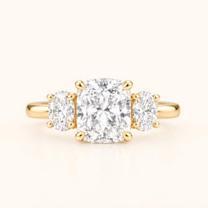 May include: A three-stone ring featuring a large cushion-cut center diamond flanked by two smaller oval-cut diamonds. The stones are set in a polished yellow gold band with a classic prong setting, showcasing a clear and brilliant design.