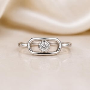 May include: A silver-toned ring featuring a horizontal, rounded rectangular frame with a single, round, clear faceted stone bezel-set in the center. The band is smooth and polished, connecting to the sides of the central geometric element.