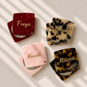May include: Four square compact mirrors with hinged lids in red, cream, pink, and brown tortoiseshell patterns. Each lid features a gold-coloured name engraved in script: Freya, Lucian, Hearth, and Mauren. The mirrors have a small metal hinge.