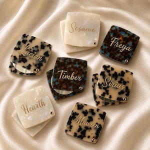 May include: A collection of square compact mirrors with hinged lids featuring various tortoiseshell and pearlescent patterns. Each lid is personalized with gold script text reading Sesame, Lucian, Freya, Timber, Sirius, Hearth, and Mauren.