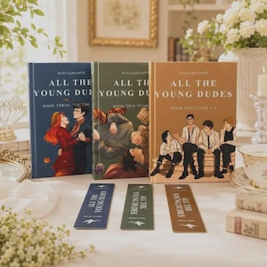 May include: Three hardback books titled All The Young Dudes by mskingbean89, arranged in a row with matching bookmarks. The blue book shows a couple dancing, the green book depicts two figures lying down, and the tan book features four seated characters.