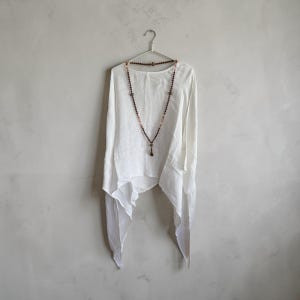 Könnte beinhalten: Ein weißes Langarmshirt mit asymmetrischem Saum und lockerer Passform. Eine lange Perlenkette mit kleinen, dunkelbraunen, kugelförmigen Perlen hängt um den Ausschnitt und liegt mittig auf dem weißen Stoff des Kleidungsstücks auf.
