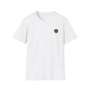 Peut inclure: Un t-shirt blanc uni à manches courtes avec un petit écusson graphique circulaire sur la poitrine gauche. Le motif circulaire présente une illustration détaillée d'un paysage de style vintage avec un texte trop petit pour être lisible.