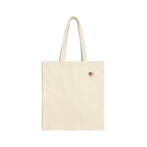 Peut inclure: Un sac fourre-tout en toile de couleur naturelle doté de deux longues anses et d'un petit écusson brodé circulaire sur le côté supérieur droit, représentant les rayures et les étoiles rouges, blanches et bleues du drapeau américain.