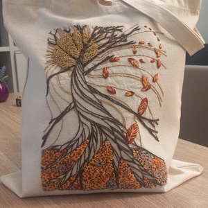 May include: A cream-colored canvas tote bag featuring an intricate embroidery design of a tree with a textured golden sun, dark brown twisted trunk, and scattered orange and red leaves, with dense, colorful foliage at the base of the tree.