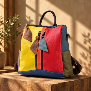 Op de afbeelding: Een veelkleurige leren rugzak met een felgeel voorpaneel, een groot rood gedeelte en blauwe en olijfgroene accenten. De tas is voorzien van twee verticale ritsvakken met leren treklipjes, een donkerbruine handgreep aan de bovenkant en schouderbanden.