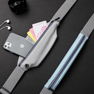 Peut inclure: Une ceinture de course gris clair avec une poche zippée contenant un smartphone et plusieurs cartes. La ceinture dispose d'une sangle élastique réglable et d'un compartiment zippé bleu clair. Le smartphone est argenté avec trois objectifs.