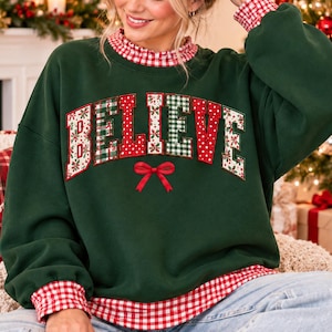 May include: A dark green long-sleeve sweatshirt featuring the word BELIEVE in large, multi-patterned block letters across the chest. Below the text is a small red bow. The collar and cuffs have a red and white gingham pattern trim.