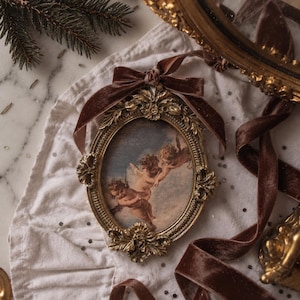 May include: An oval-shaped gold-toned picture frame featuring ornate scrollwork and floral detailing. The frame contains a print of cherubs in a blue sky and is adorned with a dark brown velvet ribbon tied into a bow at the top for hanging.