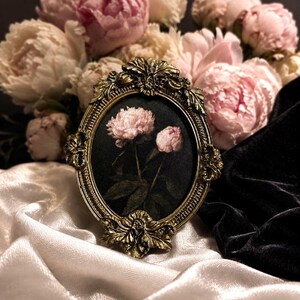 Puede incluir: Un marco de fotos ovalado con un acabado de metal dorado antiguo ornamentado que presenta detalles de hojas y volutas. El marco contiene una inserción de tela oscura que muestra una imagen impresa de dos peonías rosa claro con tallos verdes.