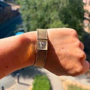 May include: A gold-tone wristwatch featuring a square face with a light-colored dial and thin dark hands. The watch has a textured, mesh-style metal band that wraps around the wrist, providing a uniform metallic appearance throughout the design.