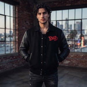 May include: A black varsity jacket featuring long black sleeves and a classic collar. The front of the jacket has a snap-button closure and displays the word "BAD" embroidered in bold red lettering on the upper left chest area.