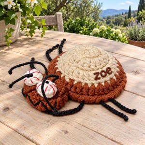 Peut inclure: Un jouet insecte en crochet composé d'un grand corps rond beige avec le prénom 'zoe' brodé en fil marron. L'insecte possède une tête marron plus petite avec deux yeux ronds blancs à coutures rouges, ainsi que des antennes et pattes en fil noir.