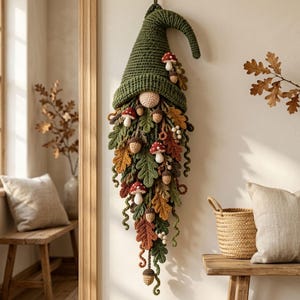 Puede incluir: Una decoración de pared de ganchillo que representa un gnomo con un sombrero alto y puntiagudo de color verde oliva. El gnomo tiene una nariz redonda de color beige y una larga barba en cascada hecha de hojas de otoño tejidas en tonos naranja, marrón y verde, adornada con pequeñas setas de sombrero rojo y bellotas marrones.
