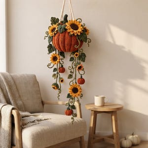 Puede incluir: Una decoración colgante de ganchillo que presenta una gran calabaza naranja central adornada con girasoles amarillos, hojas verdes y enredaderas. Calabazas naranjas más pequeñas y girasoles adicionales cuelgan de largos tallos verdes.
