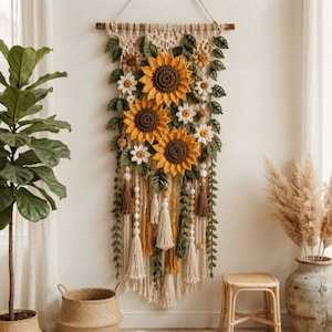 May include: A macrame wall hanging featuring three large crocheted sunflowers with brown centers and yellow petals, surrounded by smaller white crocheted flowers and green crocheted leaves. The piece includes cream-colored fringe, tassels, and wooden beads.