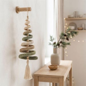 Op de afbeelding: Een spiraalvormige kerstboomversiering gemaakt van gehaakt garen in afwisselende tinten saliegroen en crème. Het ornament is voorzien van een centraal koord met houten kralen, een ophanglus en een crèmekleurig kwastje aan de onderkant.