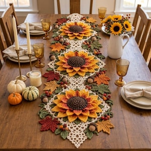 Puede incluir: Un camino de mesa de ganchillo con tres grandes girasoles tridimensionales de centros marrón oscuro y pétalos amarillo-naranja, decorado con hojas verdes, pequeñas bellotas y bayas, extendido sobre una mesa de comedor de madera con vajilla.